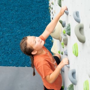 A child climbing an indoor wall with gray and green holds while reaching upward beside a blue floor mat.